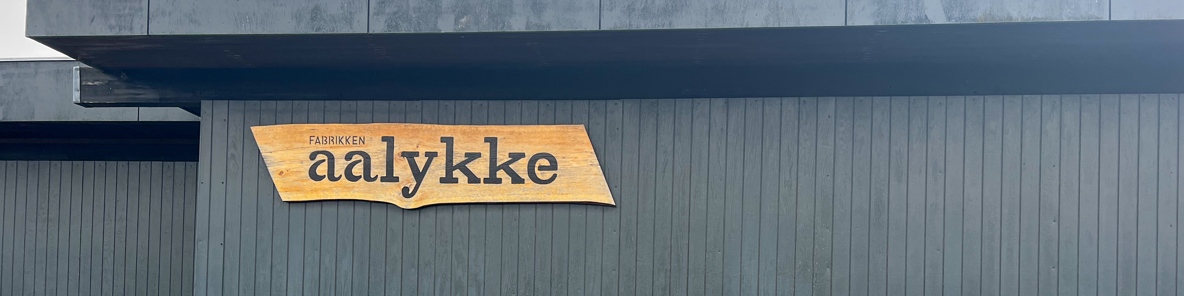 Sort træbygning med et træskilt på, hvor der står Fabrikken Aalykke på