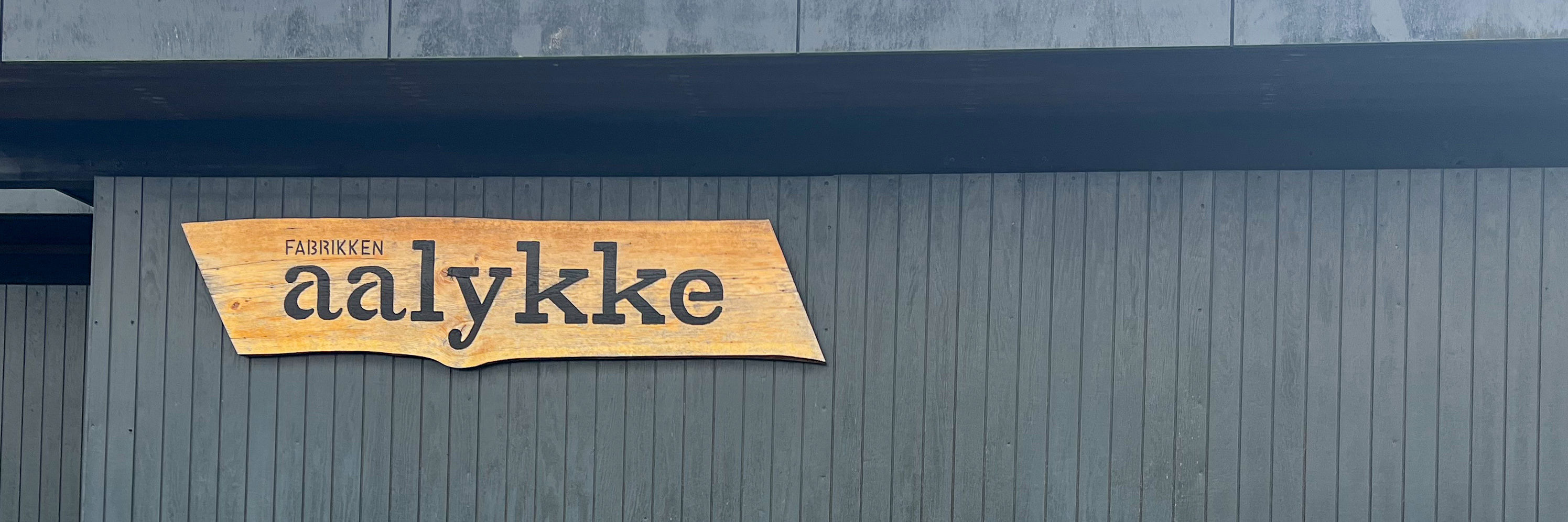 Sort træbygning med et træskilt på, hvor der står Fabrikken Aalykke på
