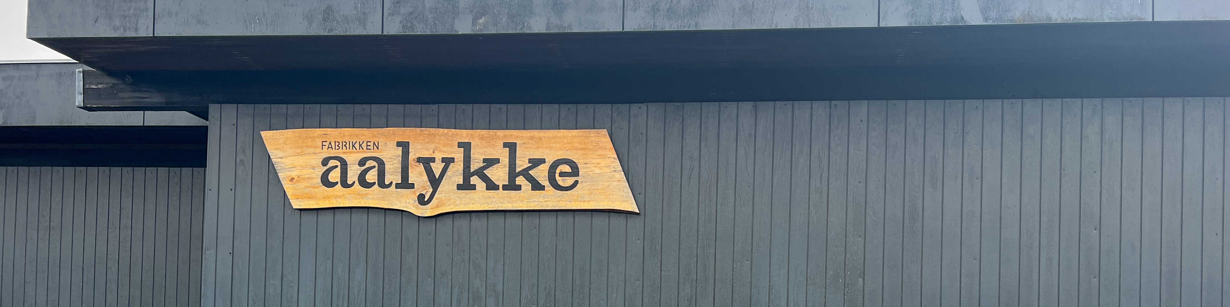 Sort træbygning med et træskilt på, hvor der står Fabrikken Aalykke på