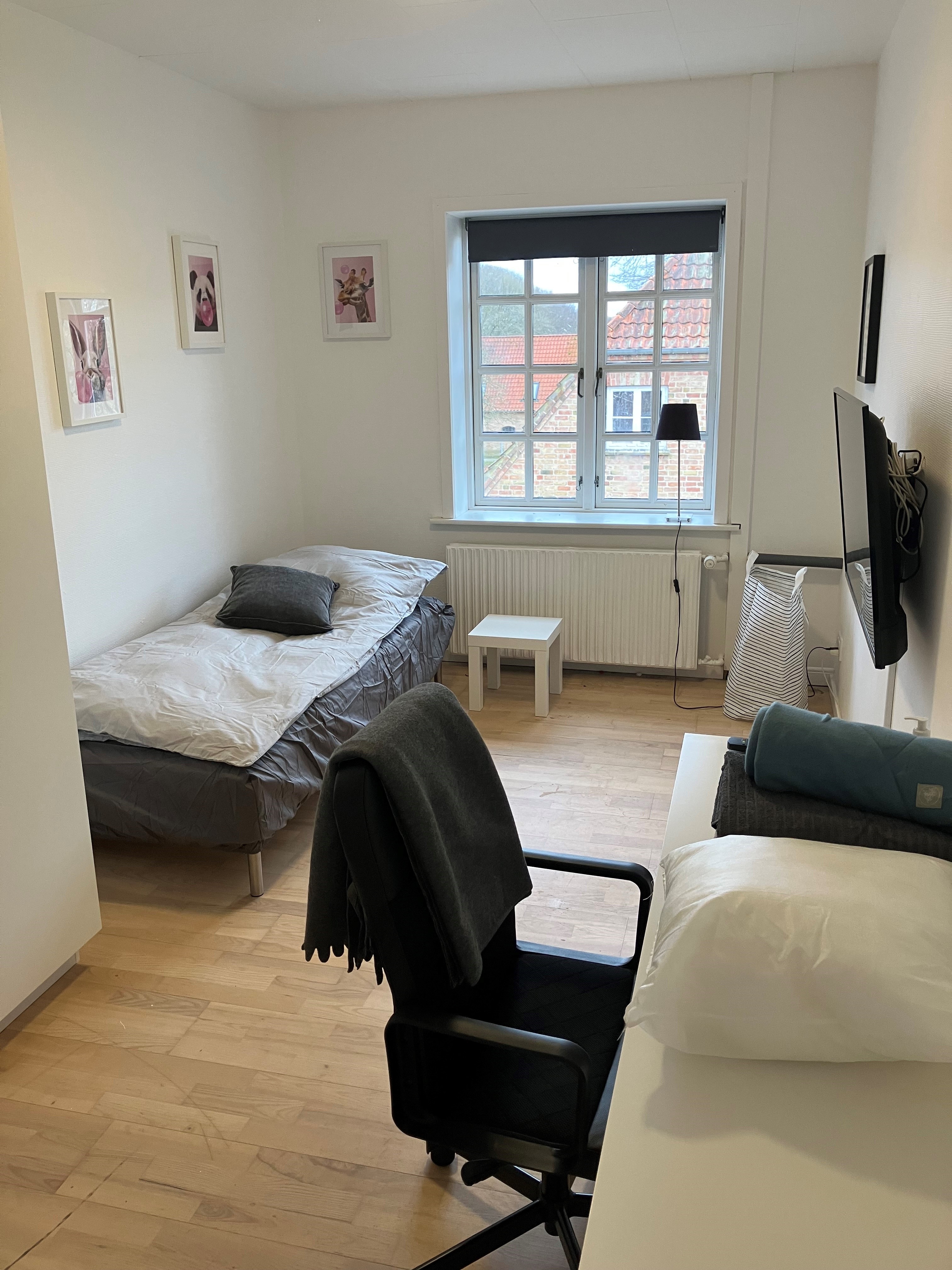 Et lyst værelse med seng, skrivebord, skrivebordsstol og tv